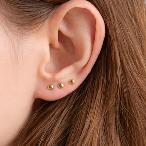 Op de afbeelding: Drie kleine, ronde, goudkleurige oorstekers zijn zichtbaar op een oor. De oorbellen zijn gelijkmatig verdeeld en lijken van metaal te zijn. Het oor is geplaatst tegen een achtergrond van bruin haar.
