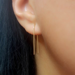 May include: A pair of gold-toned, minimalist earrings with a simple, straight design. The earrings are thin and delicate, and they hang from the earlobe.