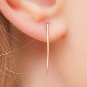 May include: A pair of gold-colored, thin, straight bar earrings. The earrings are worn in the earlobes of a person with blonde hair.
