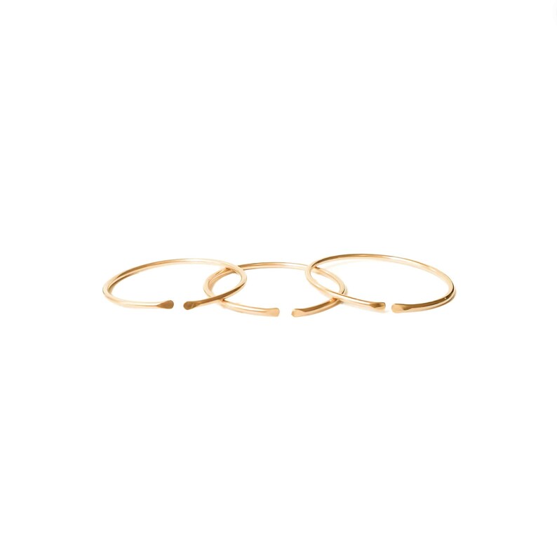 May include: Three gold-toned rings, each with a slightly different diameter, are stacked on top of each other. The rings are simple and minimalist in design, with a smooth, polished finish.