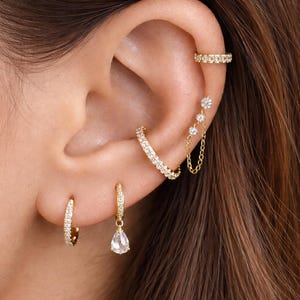 Può includere: Primo piano di un orecchio adornato con diversi gioielli color oro. L'orecchio presenta un piccolo orecchino a cerchio, un orecchino a goccia, un ear cuff curvo e un orecchino a catena con tre ciondoli a forma di fiore. Tutti i pezzi sono incastonati con piccole pietre trasparenti.