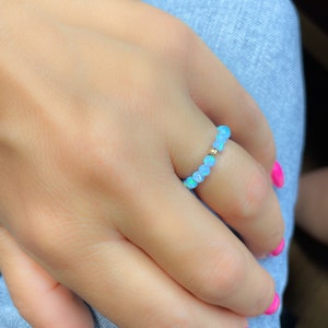 Könnte beinhalten: Ein Perlenring mit kleinen, hellblauen Opalperlen. Ein einzelnes goldenes Perle akzentuiert das Design. Der Ring wird an einem Finger getragen, mit einem hellblauen Jeans-Hintergrund.