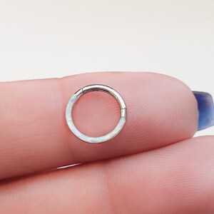 May include: A silver hoop earring with a white opal inlay. The earring is shown on a finger.