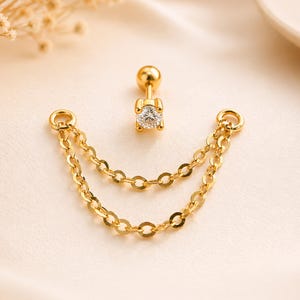 Könnte beinhalten: Goldfarbener Ohrschmuck mit einem kleinen, klaren Edelstein und einem Doppelketten-Design. Der Ohrring hat einen Stecker mit Kugelverschluss und zwei zarte Ketten. Der Schmuck wird vor einem weichen, neutralen Hintergrund präsentiert.