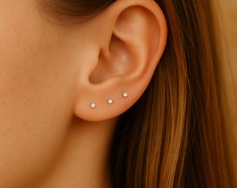 Puede incluir: Primer plano de una oreja con tres pequeños pendientes de diamantes. Los pendientes están espaciados en el lóbulo de la oreja. La piel es clara y el cabello es de color marrón cálido. Diseño minimalista.