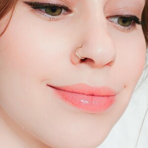 Close up of a face with green eyes and pink lips, wearing a thin polished gold hoop in one nostril.