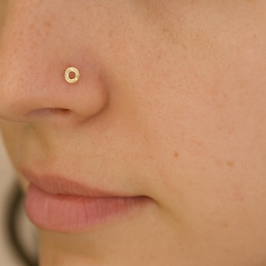 Puede incluir: Primer plano de una nariz con un pequeño piercing de nariz dorado. El piercing es de diseño simple y circular. La piel alrededor de la nariz es clara con algunas pecas. Los labios de la persona son visibles en la parte inferior de la imagen.