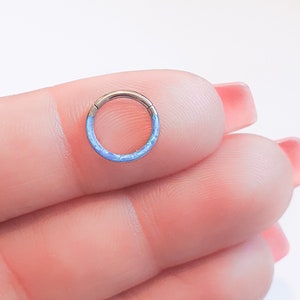 May include: A close-up of a blue opal ring, a type of body jewelry, held in a hand. The ring is made of titanium and has a smooth, polished finish.