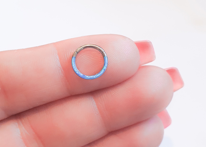 May include: A close-up of a blue opal ring, a type of body jewelry, held in a hand. The ring is made of titanium and has a smooth, polished finish.