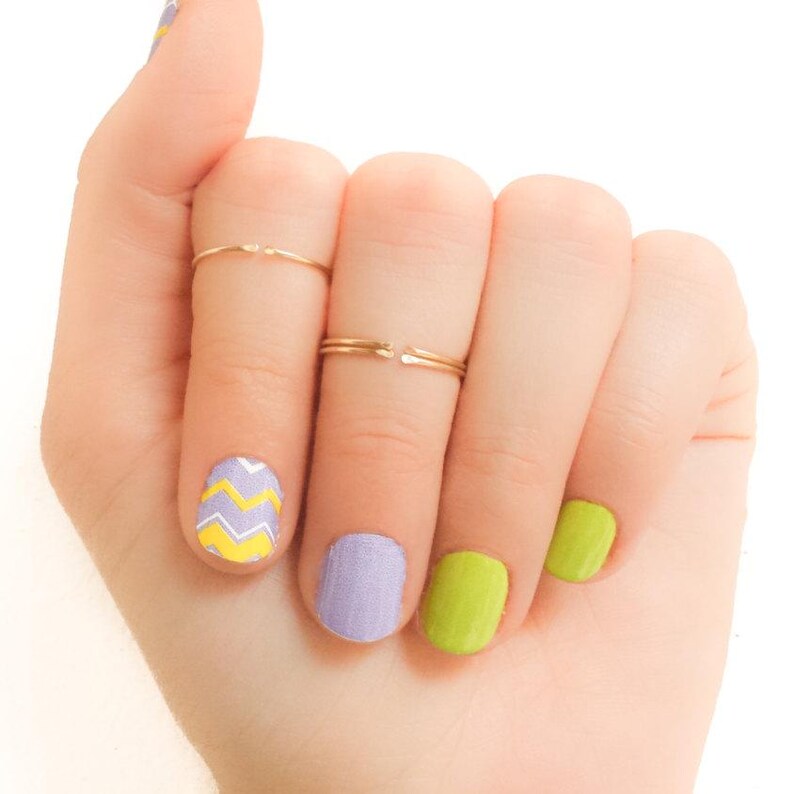 May include: A hand with four fingers showing, each with a different nail polish color. The index finger has a chevron pattern in yellow and white on a purple base. The middle finger has a solid purple polish. The ring finger has a solid lime green polish. The pinky finger has a solid lime green polish. The hand is wearing two thin gold rings on the middle finger.