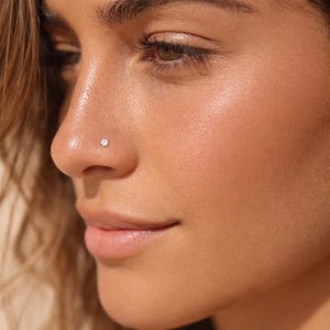 Puede incluir: Primer plano de una nariz con un pequeño piercing de nariz redondo de piedra preciosa transparente. El tono de piel es marrón claro con un brillo sutil. La atención se centra en el piercing de la nariz, destacando el delicado diseño de la joya.