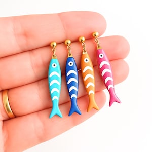 Può includere: Quattro orecchini a forma di pesce in blu, blu scuro, arancione e rosa, ciascuno con dettagli a onda bianca e borchie dorate. Gli orecchini sono tenuti in mano su sfondo bianco.
