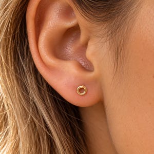 May include: A small, circular gold stud earring featuring a simple, continuous hoop design. The earring is positioned in the earlobe, showcasing a polished, metallic finish and a minimalist, open-ring shape that sits flush against the skin.