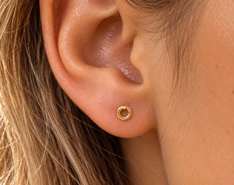 May include: A small, circular gold stud earring featuring a simple, continuous hoop design. The earring is positioned in the earlobe, showcasing a polished, metallic finish and a minimalist, open-ring shape that sits flush against the skin.