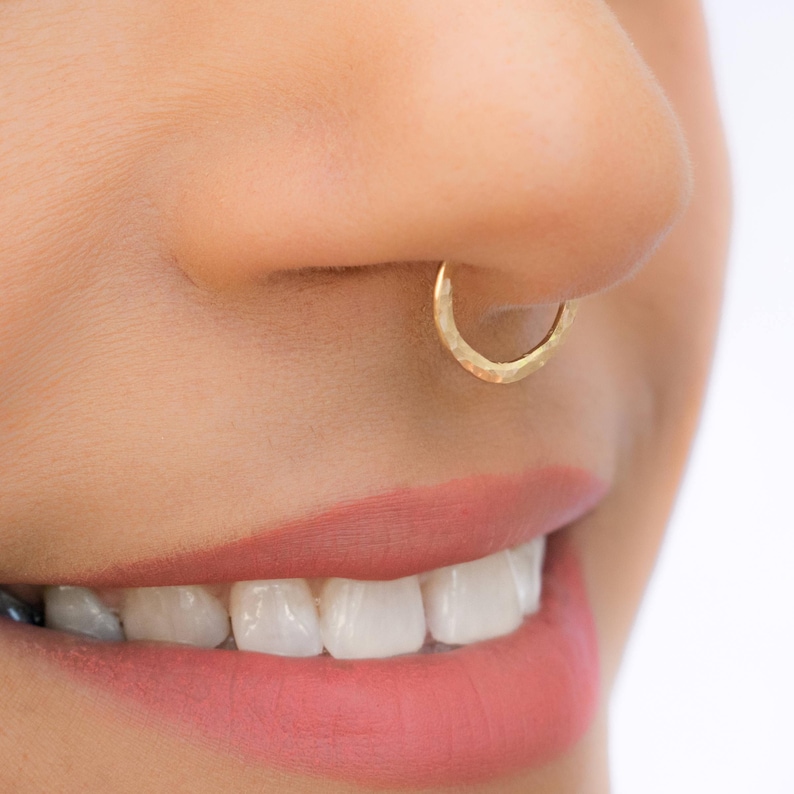 Side view of a smiling face with a hammered gold hoop worn in the septum, pink lips and white teeth below.