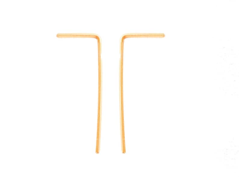May include: A pair of gold-toned earrings with a simple, minimalist design. Each earring is a thin, straight piece of metal that is bent at a right angle to form a small, angled shape.