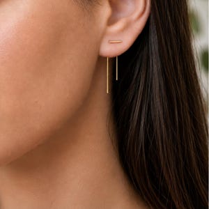 Puede incluir: Primer plano de una oreja con pendientes dorados. Los pendientes presentan una pequeña barra horizontal en el lóbulo, con dos delicadas barras rectas doradas colgando. La joya tiene un diseño minimalista.