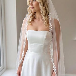Könnte beinhalten: Ein weißes, trägerloses Brautkleid mit fließendem Rock. Ein durchsichtiger Schleier mit zarter, blattartiger Stickerei umrahmt das Gesicht und die Schultern. Die Braut schaut aus dem Fenster.