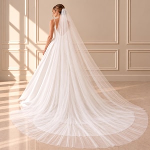 May include: A floor-length white wedding veil made of sheer, lightweight tulle fabric. The veil is attached at the crown and cascades down the back, extending into a long, wide train that spreads across the floor in a circular shape.