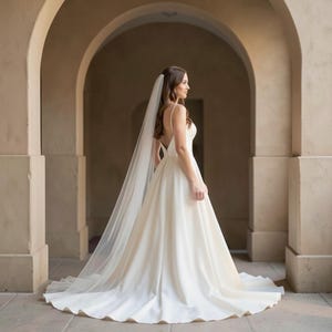 May include: A white bridal gown with a flowing skirt and spaghetti straps. A long, sheer veil trails down the back. The dress is worn in an arched walkway with a stone floor.