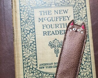 May include: A brown leather bookmark with a cat face embroidered on it. The bookmark is sitting on top of a vintage book titled "The New McGuffey Fourth Reader".