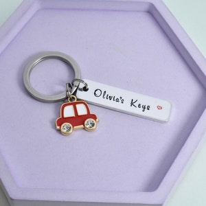May include: A red car keyring with a white roof and gold accents. The keyring has a silver ring and a white tag with the text "Olivia's Keys" and a small red heart.