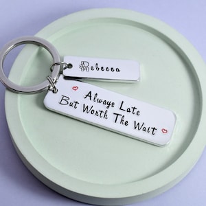 May include: Silver keyring with two rectangular tags. The top tag says "Rebecca" and the bottom tag says "Always Late But Worth The Wait" with a small heart on each side of the text.