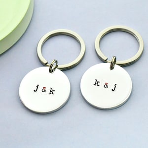 May include: Two silver keyrings with circular pendants. Each pendant has a silver keyring and features initials with a small red heart. One keyring says "j & k", the other says "k & j".