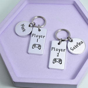 May include: Two silver keychains with engraved names, Mark and Sabrina. One keychain has a silver tag that says "Player 1" with a video game controller icon. The other keychain has a silver tag that says "Player 2" with a video game controller icon.
