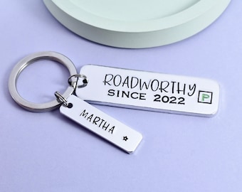 Puede incluir: Llavero de plata con dos etiquetas. La etiqueta más grande dice "Roadworthy Since 2022" con una señal de estacionamiento verde. La etiqueta más pequeña dice "Martha" con una estrella.