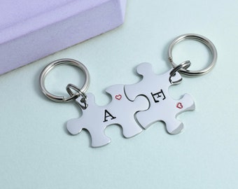 May include: Two silver puzzle piece keychains with the letters A and E engraved on them. Each piece has a small red heart engraved on it.