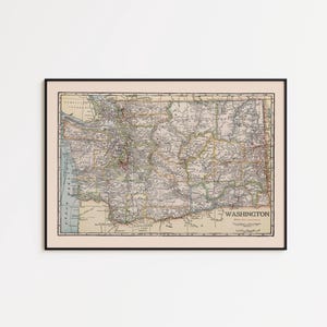 May include: Framed vintage map of Washington state, featuring detailed geographical information. The map is in a black frame with a cream-colored border, and the map itself has a beige background with various colors highlighting different regions and features. The word "WASHINGTON" is visible.