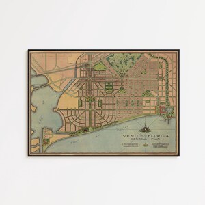 May include: A framed vintage map titled Venice Florida General Plan, featuring a detailed grid of streets, green spaces, and coastal areas in muted tones of tan, green, and blue, enclosed within a thin black frame against a plain white wall.