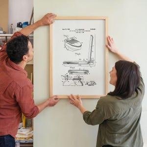 May include: A framed patent print featuring technical drawings of a toilet design. The print is in black and white, with detailed diagrams and text. The frame is made of light-colored wood. Two people are holding the framed print.