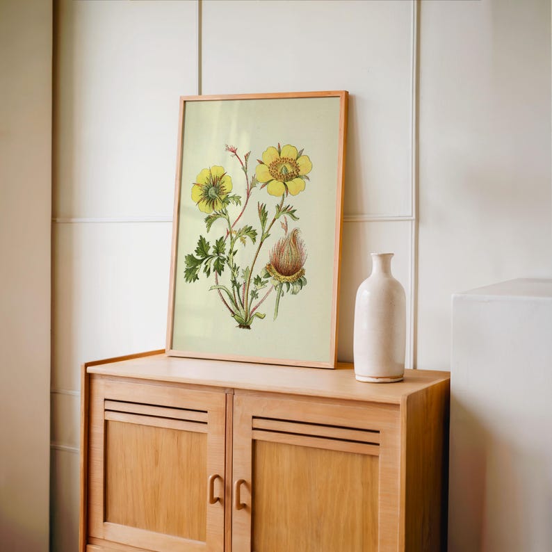 May include: A framed botanical print featuring yellow flowers and green leaves. The artwork is displayed on a light-colored cabinet with a white vase. The print has a light-colored wooden frame.