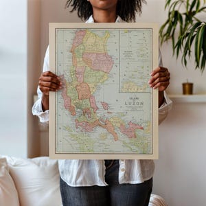 Può includere: Una mappa vintage incorniciata dell'isola di Luzon, Filippine, con un bordo beige. La mappa presenta vari colori per indicare le regioni e include il testo "Island of Luzon". La mappa è tenuta da una persona.