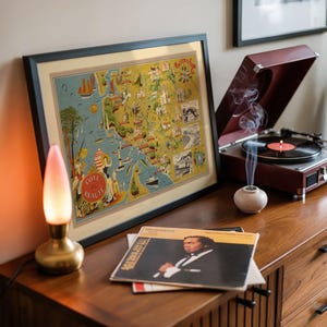 May include: A framed vintage-style map print titled Cote de Beaute featuring illustrations of coastal landmarks, a brass lamp with a pink-tinted glass shade, a maroon record player with a spinning vinyl record, and a stack of vinyl record sleeves.