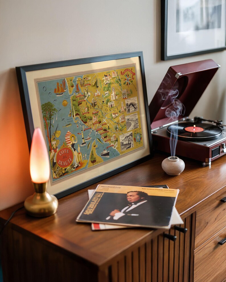 May include: A framed vintage-style map print titled Cote de Beaute featuring illustrations of coastal landmarks, a brass lamp with a pink-tinted glass shade, a maroon record player with a spinning vinyl record, and a stack of vinyl record sleeves.