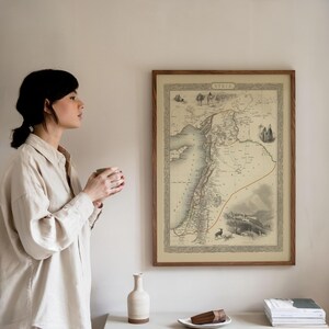 May include: A framed vintage map of Syria, featuring detailed geographical elements such as coastlines and rivers. The map is presented in a wooden frame and hangs on a wall. The map has a beige background with black and red lines.
