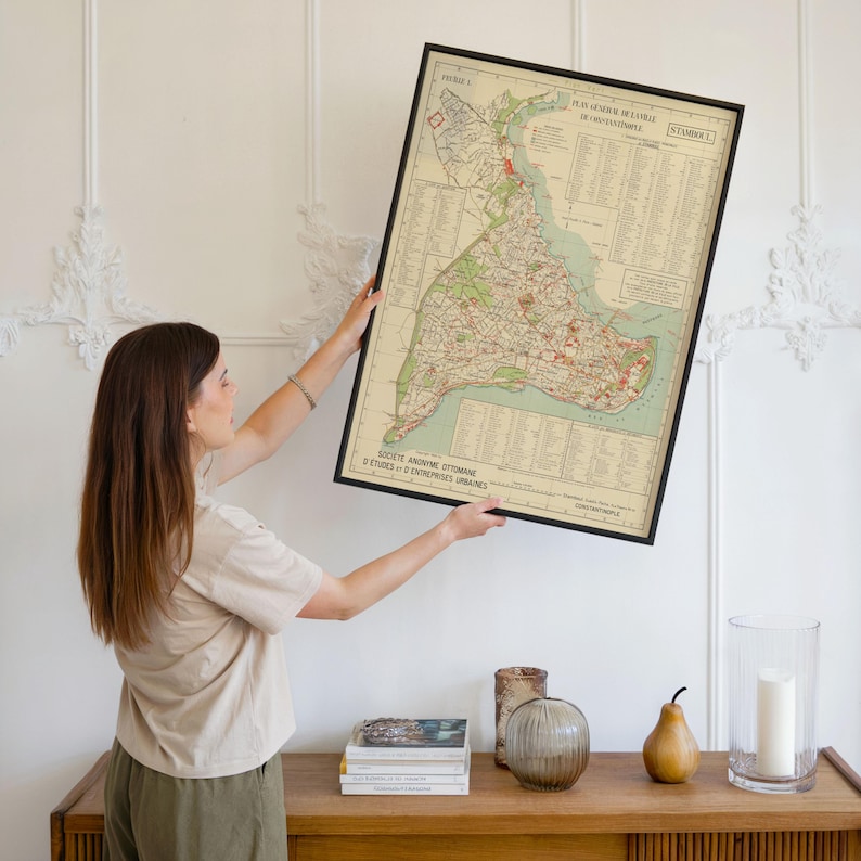 May include: A framed vintage map of Istanbul, Turkey, with detailed city planning information. The map features a black frame and is held against a white wall. The map includes text in French. The overall color palette is beige, green, and red.