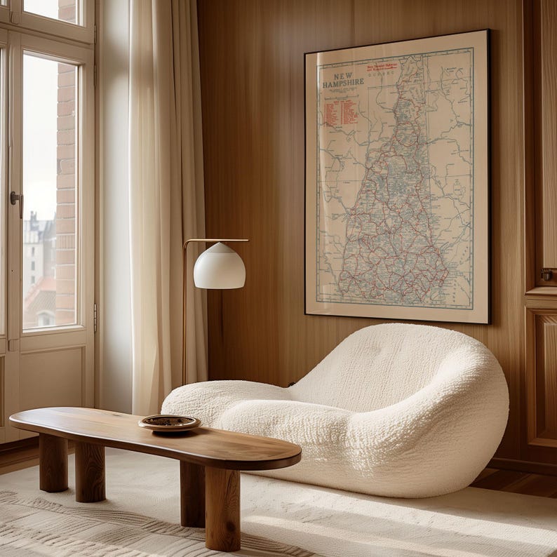 May include: A cozy living room scene featuring a cream-colored, curved sofa and a wooden coffee table. A framed map of New Hampshire hangs on the wood-paneled wall. A white lamp with a gold arm provides soft lighting. A rug covers the floor.