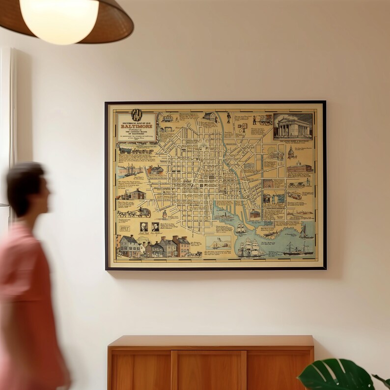May include: A framed historical map of Baltimore, Maryland, featuring detailed street layouts, illustrations of ships, buildings, and horse-drawn carriages, with text labels and a beige background, displayed in a black frame on a plain wall.