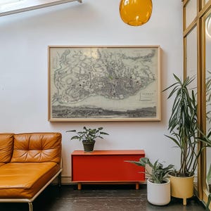 May include: A framed vintage map of Lisbon hangs on a white wall above a rectangular orange cabinet. To the left is a tan leather sofa, and to the right are two potted green plants in white and yellow containers, with an amber glass pendant light above.