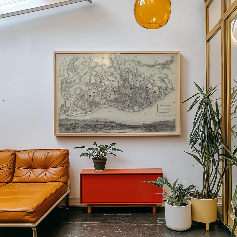 May include: A framed vintage map of Lisbon hangs on a white wall above a rectangular orange cabinet. To the left is a tan leather sofa, and to the right are two potted green plants in white and yellow containers, with an amber glass pendant light above.
