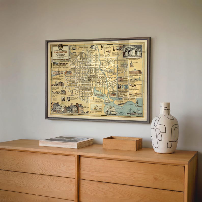 May include: A framed historical map of Baltimore, Maryland, featuring a grid of city streets, illustrations of local landmarks, and ships in the harbor. The map has a cream-colored background with black ink details and is displayed in a dark frame.