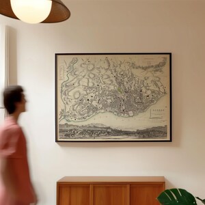 May include: A framed vintage-style map of Lisbon, Portugal, displayed on a plain wall. The map features detailed black and white cartographic illustrations of the city layout, including street grids, topography, and a panoramic view of the harbour.