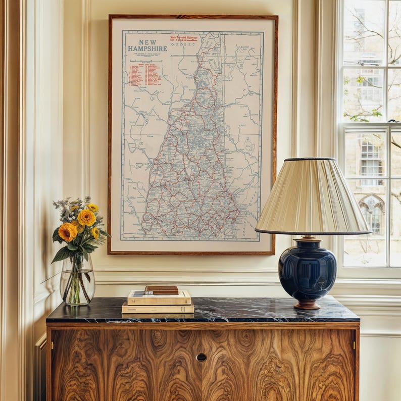 May include: A framed vintage map of New Hampshire hangs above a wooden cabinet. A navy blue lamp with a cream-colored shade sits on the cabinet. A vase of yellow flowers and a stack of books complete the decor.