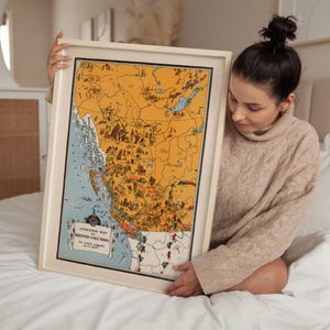 May include: A framed illustrated map titled Animated Map of British Columbia, featuring a yellow landmass with various small drawings of animals, people, and landmarks, and a blue coastal area, all enclosed in a light-colored wooden frame.