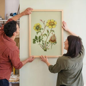 May include: A framed botanical print featuring yellow flowers and green leaves. The artwork has a light green background and is encased in a natural wood frame. The print is being held up by two people.