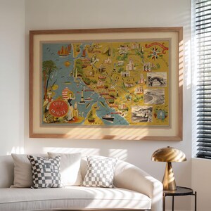 May include: A framed vintage-style map print titled Cote de Beaute, featuring a yellow and blue illustrated coastal landscape with ships, lighthouses, and small inset black and white photographs, enclosed in a light-toned wooden frame.